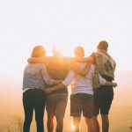 four person hands wrap around shoulders while looking at sunset