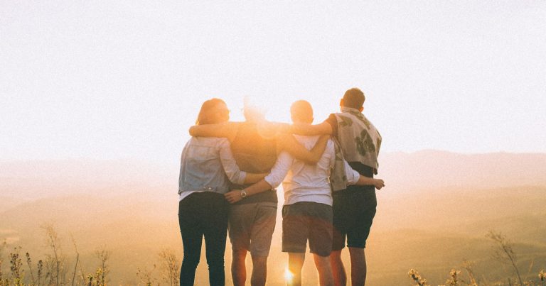 four person hands wrap around shoulders while looking at sunset